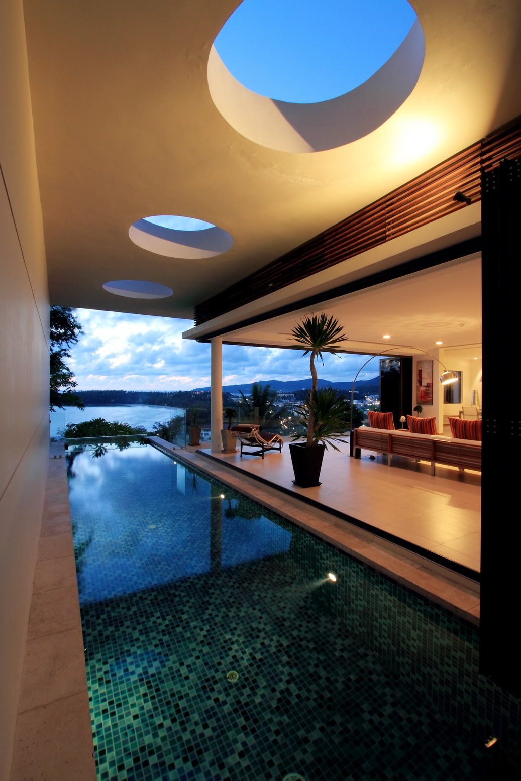 The image displays a private pool surrounded by a sleek patio area, with sliding glass doors opening to a modern living space. Unique circular skylights offer ambient light, while views of the Andaman Sea are framed by lush greenery and a calming evening sky.