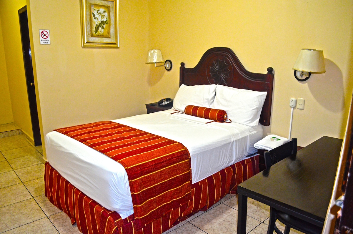 A simple room features a spacious bed with a red and white striped throw blanket. Soft lighting is provided by bedside lamps. A desk is positioned beside the bed, and a door leads to a hallway. The walls are painted in a neutral tone.