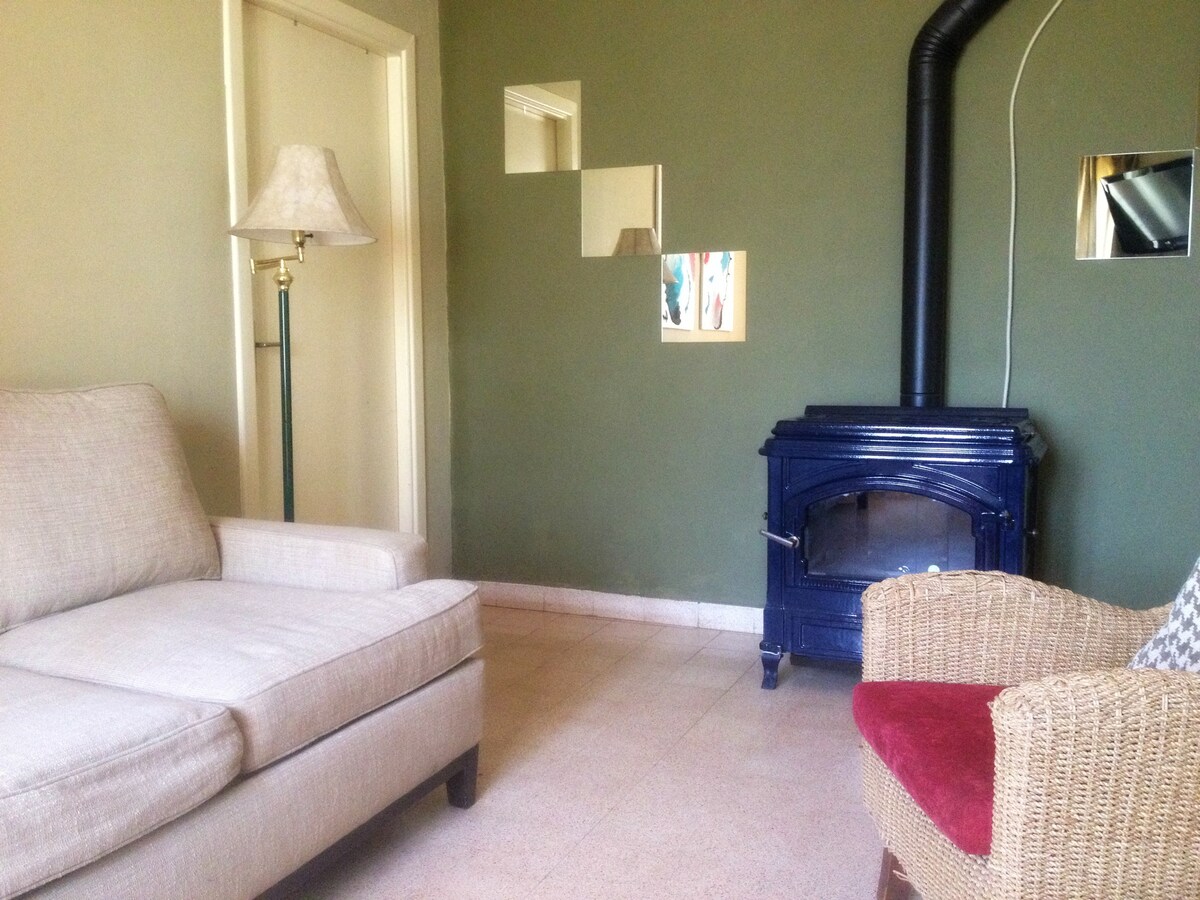 A cozy living room is highlighted by a comfortable couch and a patterned wicker armchair. A kerosene stove provides warmth, while mirrors reflect light across the green walls. A floor lamp stands beside the couch, and natural light from a nearby window softly illuminates the space.