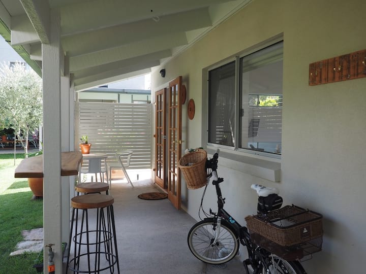  The image shows a covered outdoor patio area with a small seating arrangement featuring a table and two chairs. The space includes a wooden bar counter with high stools along the wall, and a bicycle with a wicker basket is parked against the house. French doors lead inside, and a modern privacy screen provides separation between the patio and the yard, which is filled with greenery. The overall atmosphere is serene and functional, with a blend of practicality and outdoor living