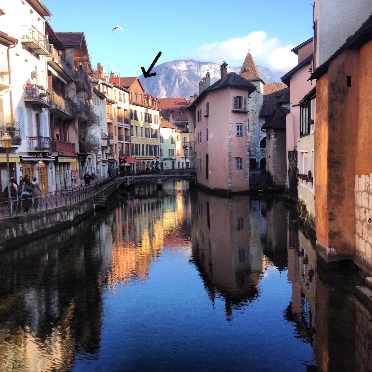 A serene canal reflects colorful buildings lining its banks, showcasing a blend of architectural styles. In the background, a mountain is visible under a clear sky, adding to the tranquil atmosphere. The image captures the essence of the old town with its picturesque waterside setting.
