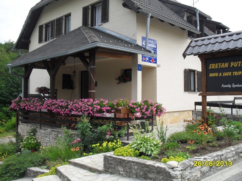 A welcoming entrance to a multi-level house is highlighted by a charming front porch adorned with vibrant pink flowers and various greenery. The structure features a sloped roof and wooden accents. A sign indicating the property name stands nearby amidst the blooming garden.