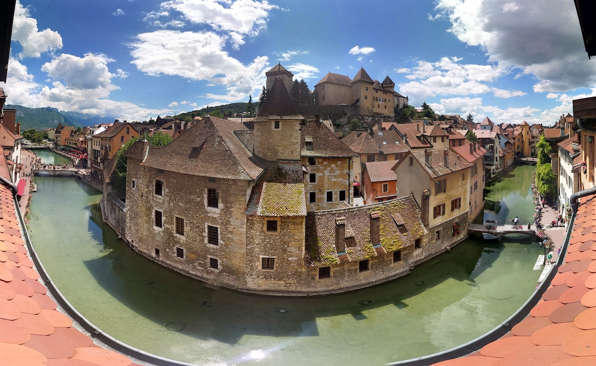 A panoramic view of a historic town is captured, featuring charming stone buildings with sloped roofs along a winding canal. Lush greenery complements the scene, while a distant castle sits atop a hill, under a sky scattered with clouds.