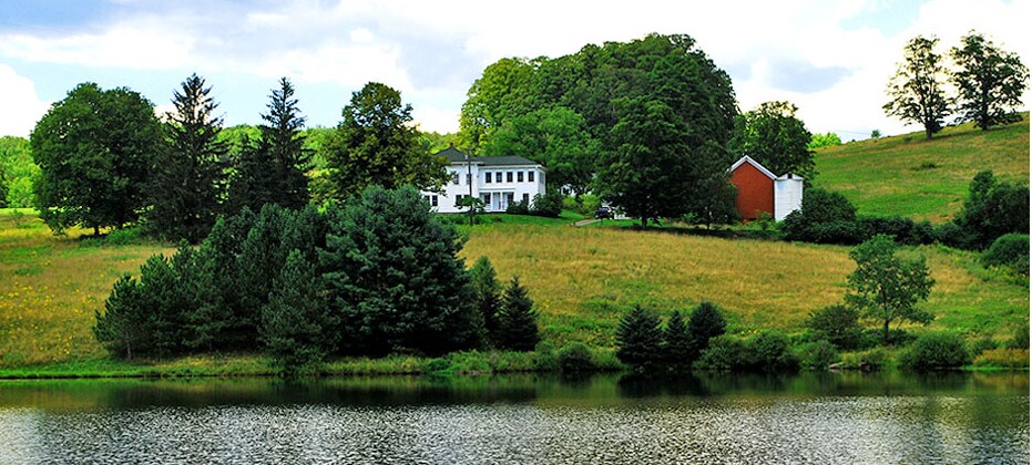 The farmhouse is visible on a gently rolling landscape, surrounded by vibrant greenery. A lake reflects the structure and the lush trees lining its shore, creating a serene natural setting. The area is characterized by tranquil open spaces and a mix of trees and grass.