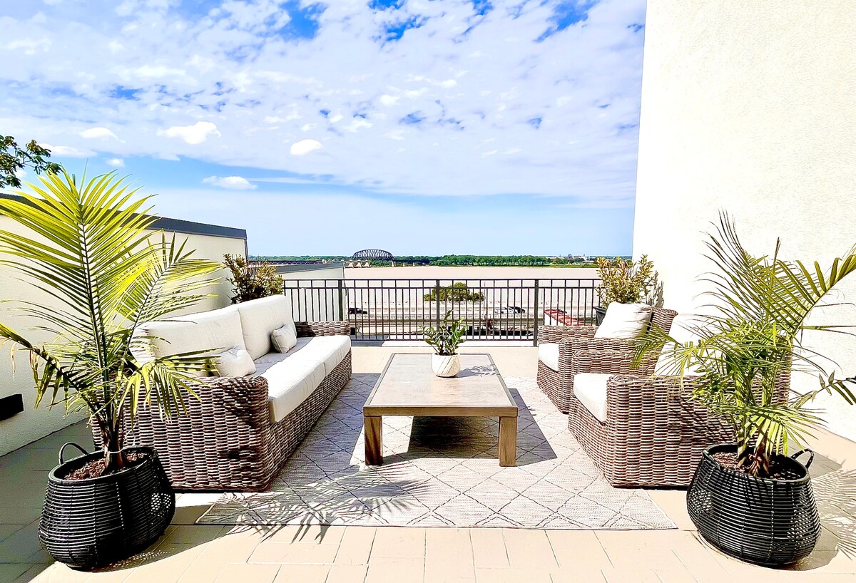 A spacious rooftop patio is highlighted by stylish furniture, including a large sectional and coffee table. Potted plants add greenery, while a scenic river view stretches in the background beneath a clear blue sky filled with light clouds.
