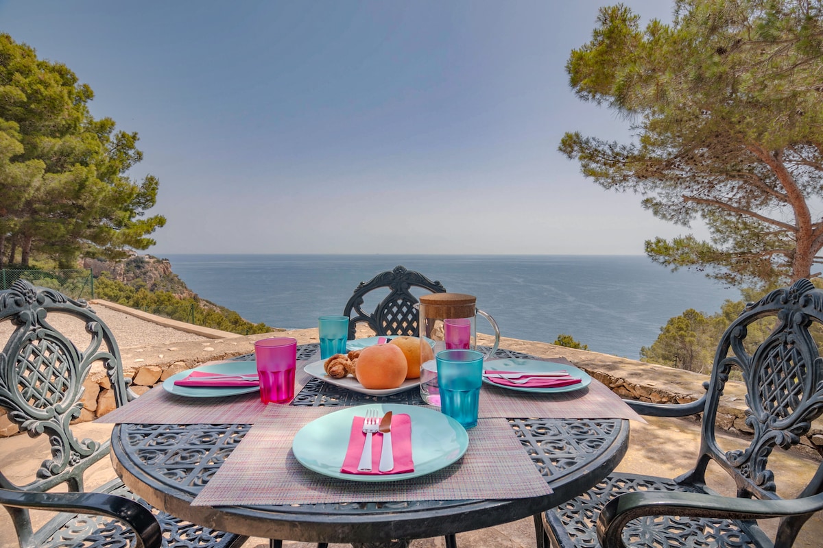 An outdoor dining table is set for a meal, featuring round plates, glasses, and cutlery. The table is surrounded by wrought iron chairs, with a backdrop of the Mediterranean Sea visible in the distance. Trees provide a natural frame to the serene landscape.