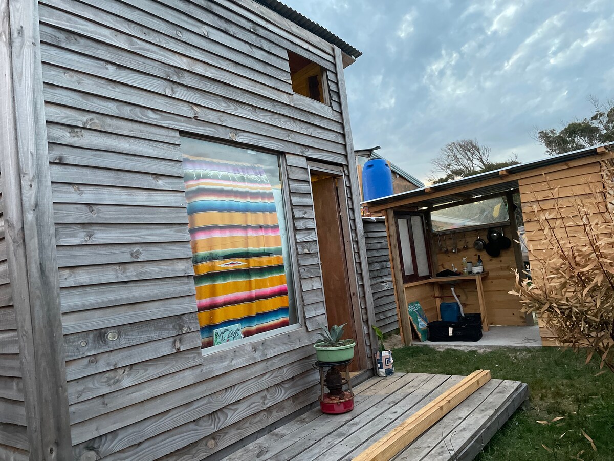 A wooden ranch-style exterior is visible, featuring a colorful striped curtain in a large window. Nearby, an outdoor kitchen area is partially sheltered, with utensils and equipment arranged neatly. Green grass surrounds the structure, contributing to a rustic yet homey atmosphere.
