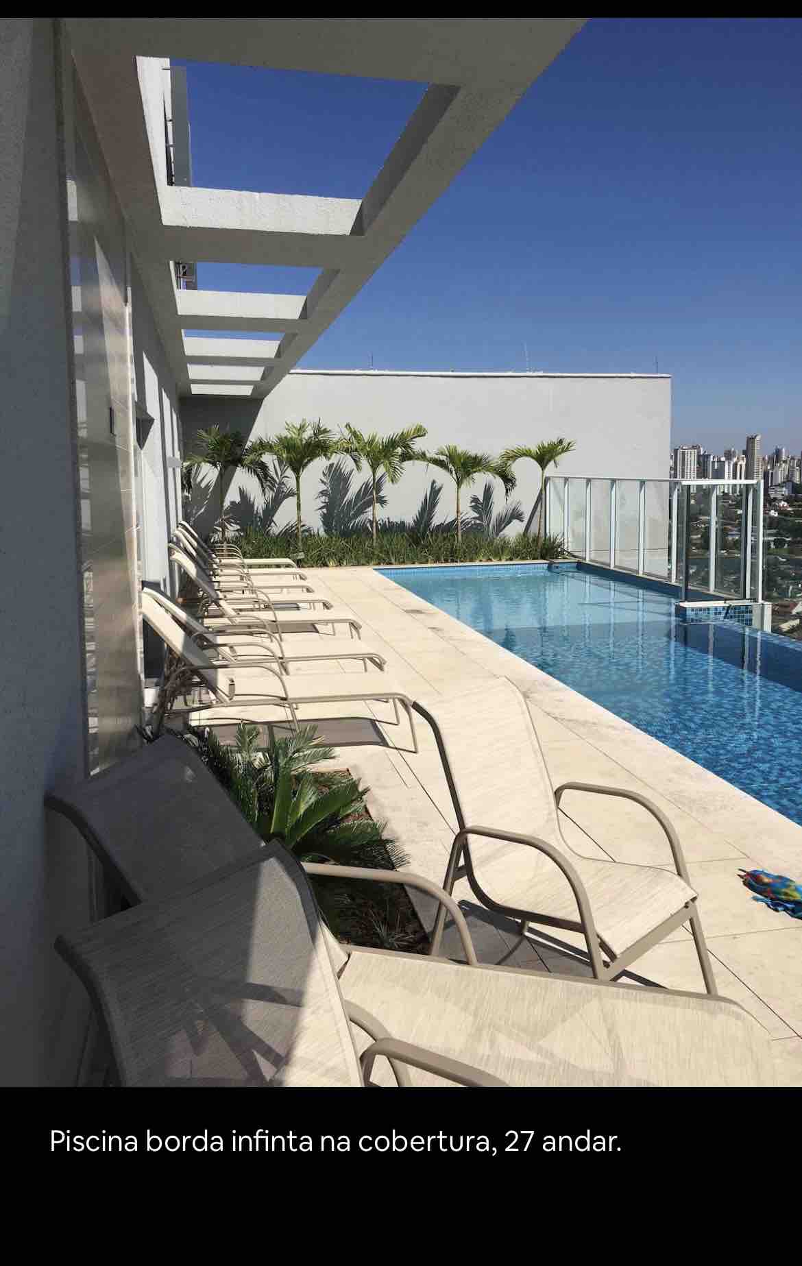 An infinity pool is situated on the rooftop, offering panoramic city views. Several lounge chairs line the poolside, surrounded by greenery and neatly tiled decking. The calm water reflects the clear blue sky, creating a serene outdoor setting.