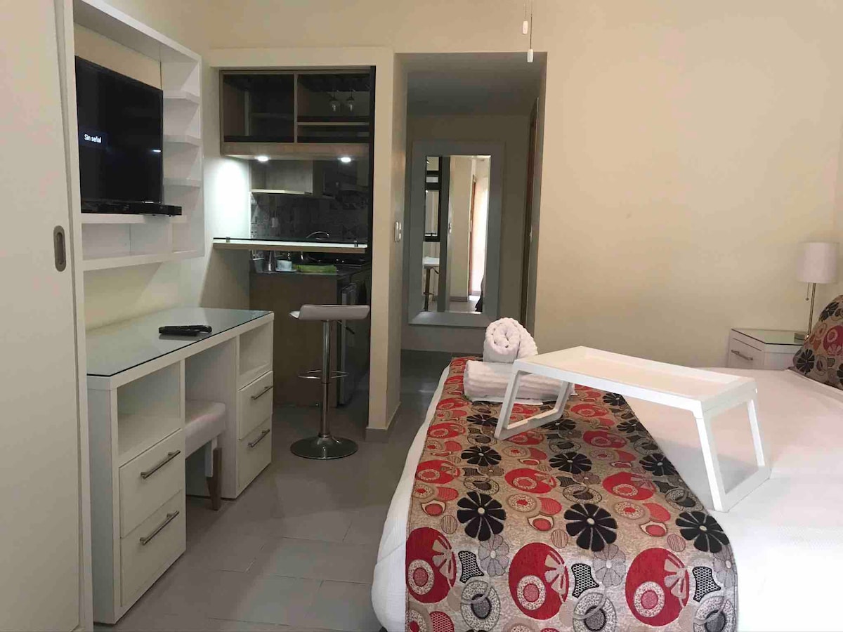 A modern suite with a spacious bed adorned in a colorful patterned bedspread. A small kitchenette is integrated in the background, featuring a bar-style seating area. A television is mounted on the wall, while a desk and a white serving tray are positioned nearby.