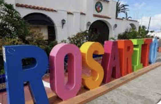 A large, colorful sign spelling 'Rosarito' stands prominently outside a building. The letters are painted in bright hues of orange, pink, yellow, and blue, set against a backdrop of greenery and architecture typical of the area.