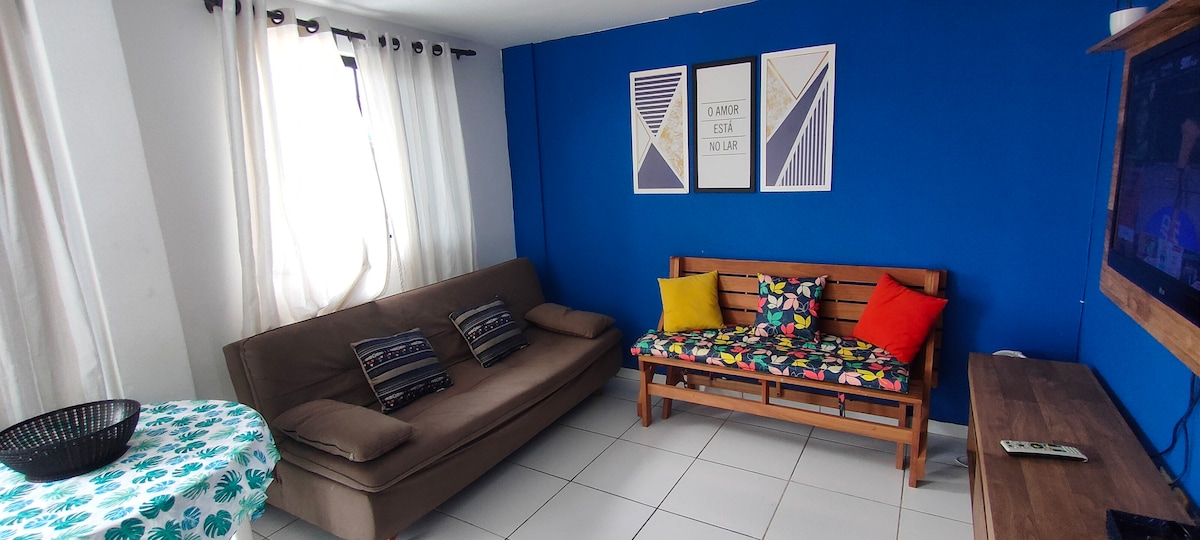 A living area features a brown sofa with soft cushions and a wooden bench adorned with colorful pillows. A round table with a floral pattern is visible, along with a television mounted on the wall. Natural light filters through sheer curtains.