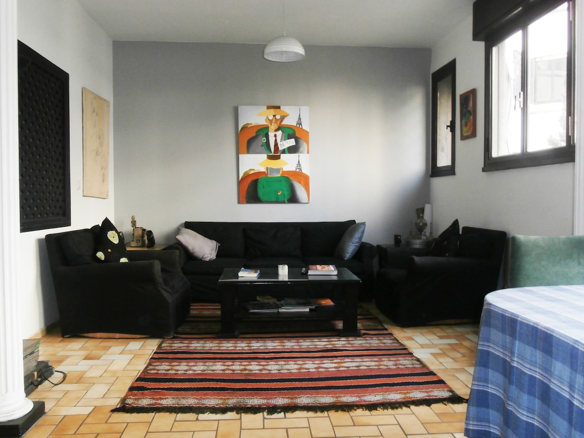 A cozy living area showcases a dark-colored sofa with assorted cushions, complemented by a central coffee table. A vibrant area rug lies beneath, while a large artwork adorns the wall. Two windows provide natural light, enhancing the spacious feel of the room.