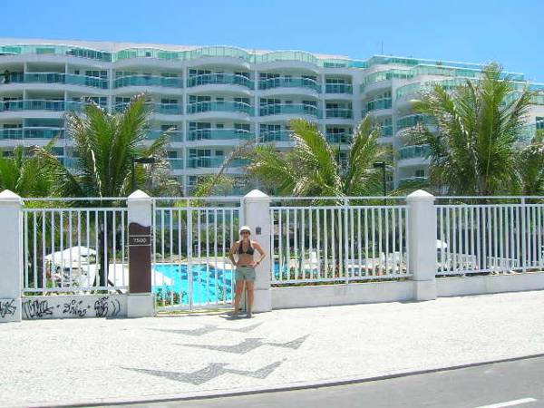 A modern condominium building is visible beyond a white gated entrance, framed by lush palm trees. A swimming pool is partially seen, indicating an outdoor recreation area. The blue sky enhances the welcoming atmosphere of the property.
