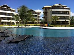 A spacious outdoor pool is framed by palm trees and modern resort-style buildings. Comfortable lounge chairs are positioned along the pool's edge, inviting relaxation. The clear blue sky enhances the inviting atmosphere, while vibrant landscaping adds to the resort's appeal.