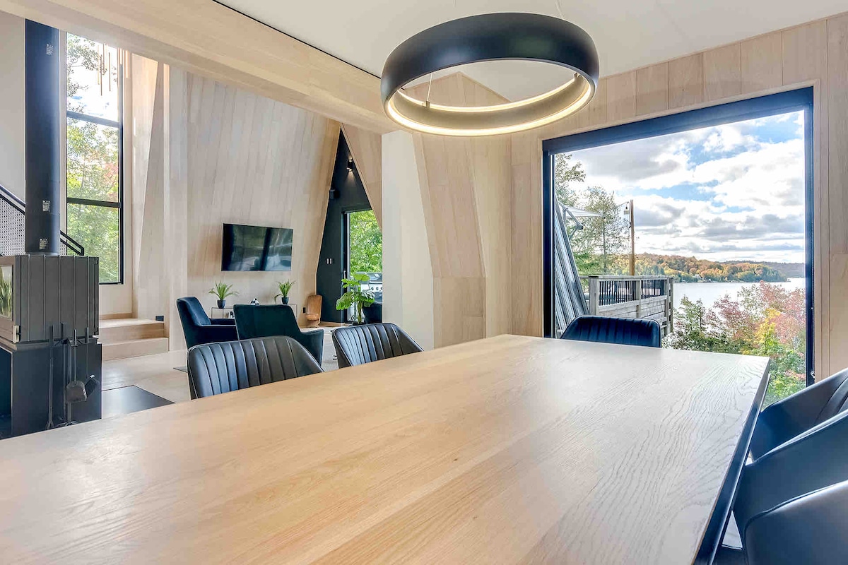 A spacious dining area features a light wooden table surrounded by black chairs. Large windows reveal views of the lake and surrounding nature. The living area is partially visible, showcasing a black television and angled walls that create a modern aesthetic.