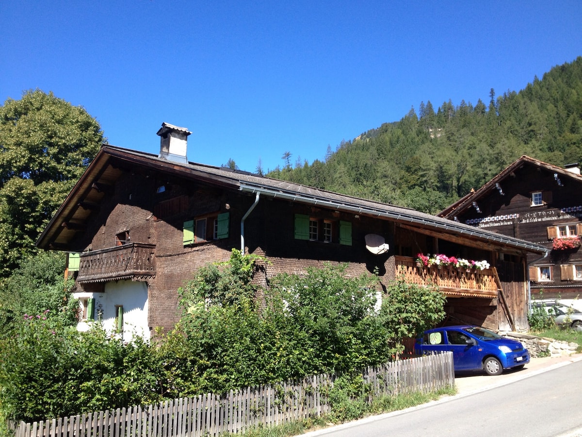 The charming wooden exterior of the accommodation is framed by lush greenery and vibrant flowers adorning the balcony. The building features traditional mountain architecture with a sloped roof and wooden shutters. A quiet road runs alongside the property, with a blue car parked nearby.