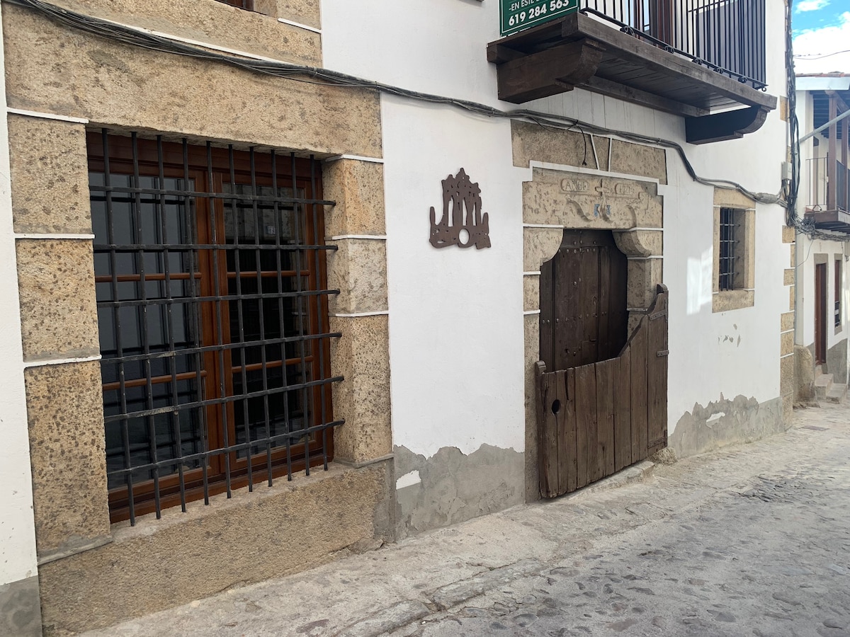The exterior of a traditional stone building is visible, showcasing a rugged texture with wooden window grilles. A weathered wooden door is centered, surrounded by whitewashed walls. The cobblestone pathway adds to the charm of this inviting street scene.