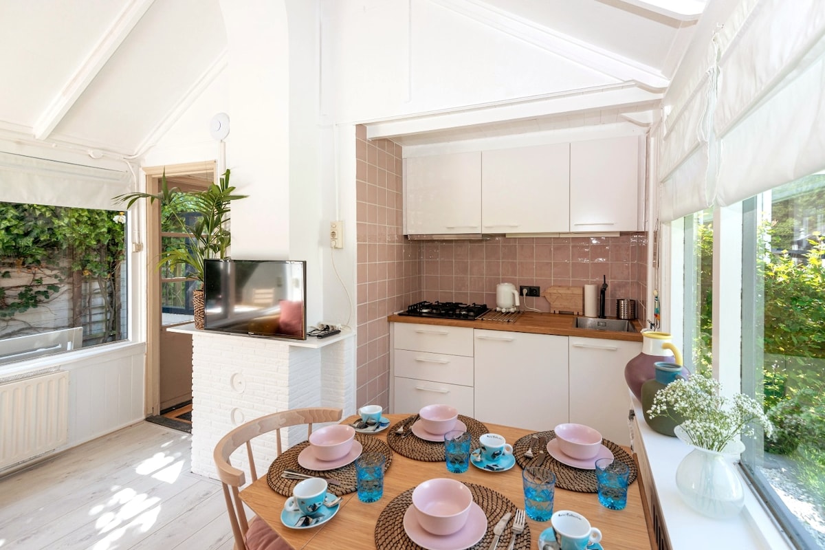 A bright dining area features a round wooden table set for six, surrounded by woven placemats and vibrant turquoise glassware. The kitchen is seen in the background, highlighting modern cabinetry and a white counter. Natural light filters through large windows, illuminating the space.