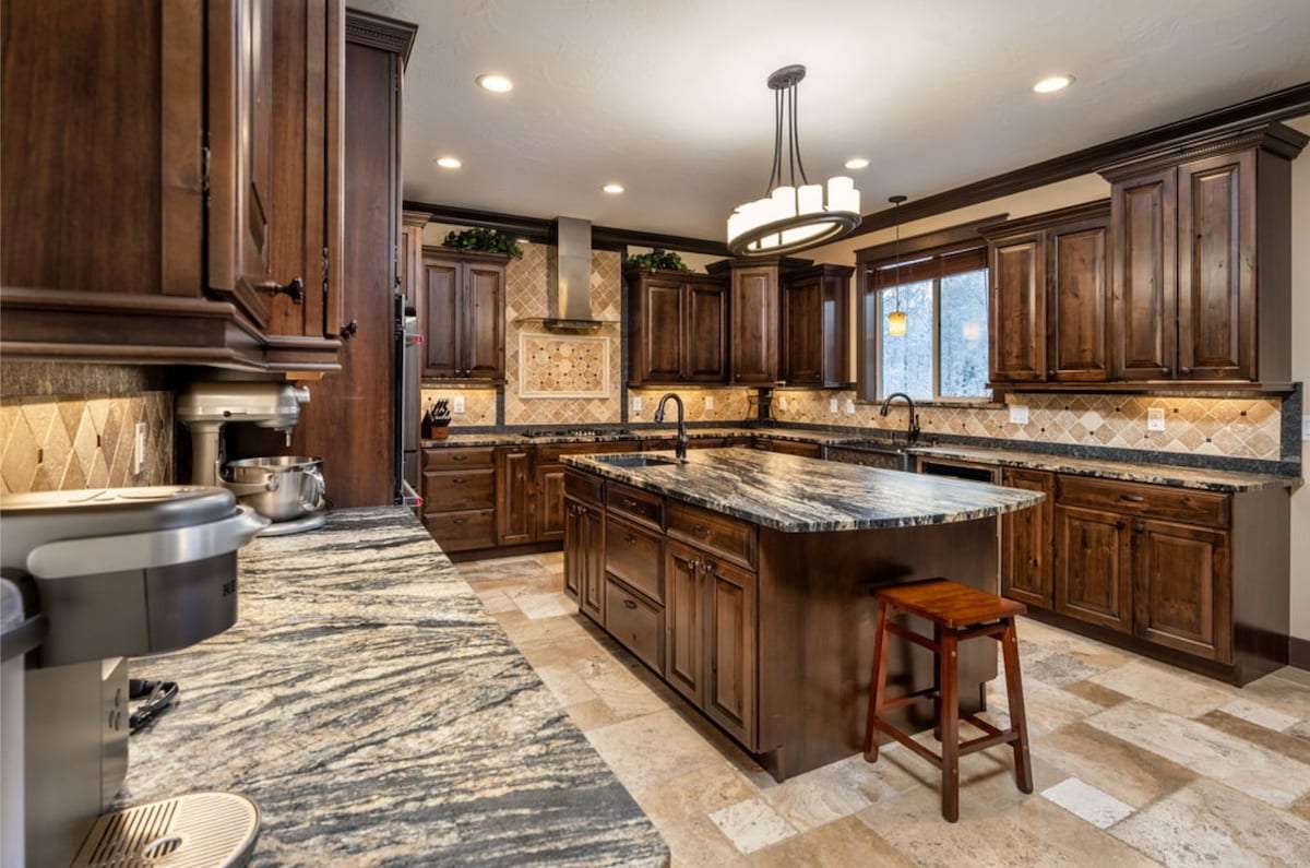 A spacious kitchen features rich wooden cabinetry and a large island with a granite countertop. A bar stool is positioned at the island, while tile flooring complements the elegant design. Natural light flows through the window, enhancing the warm tones of the wood and stone.