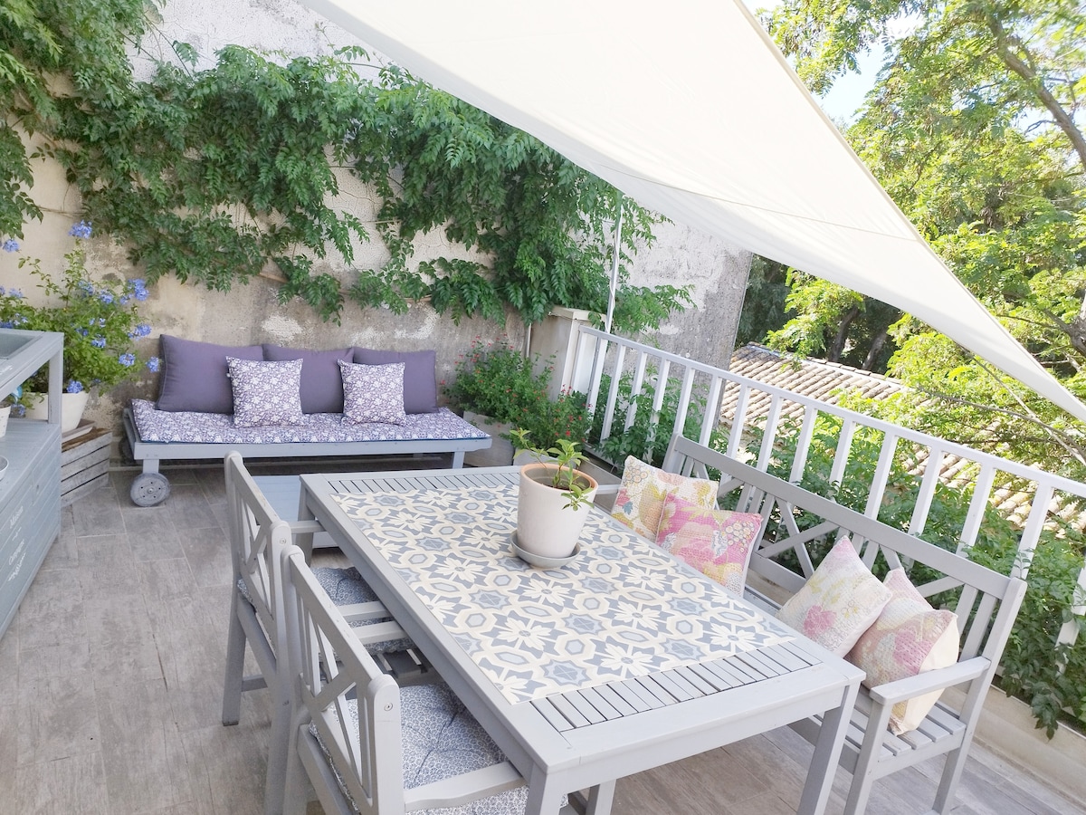 A serene terrace area features a handcrafted wooden seating arrangement, complemented by a sofa adorned with decorative cushions. A patterned table sits at the center, surrounded by additional seating. Lush greenery adds a natural touch, creating a cozy, private outdoor escape.