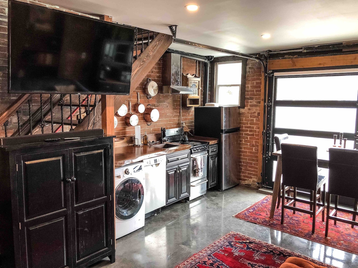 A functional living space features a kitchen area with modern appliances, including a gas stove and washing machine. Dark cabinetry complements the exposed brick walls. A television is mounted on the wall, and a dining table is placed near a large window, offering ample natural light.