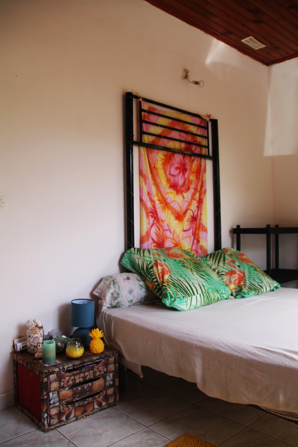 A cozy bedroom is highlighted with a colorful floral wall hanging above a spacious bed. Large green pillows rest on the bed, while a small decorated table holds tropical-themed decor. Soft lighting from a bedside lamp creates a warm, inviting space.