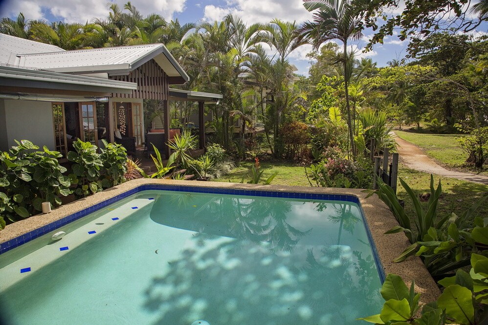 A private pool glistens under the sun surrounded by lush greenery, showcasing a blend of palm trees and tropical plants. The inviting outdoor space leads to the entrance of a cottage, creating a serene atmosphere that emphasizes relaxation.