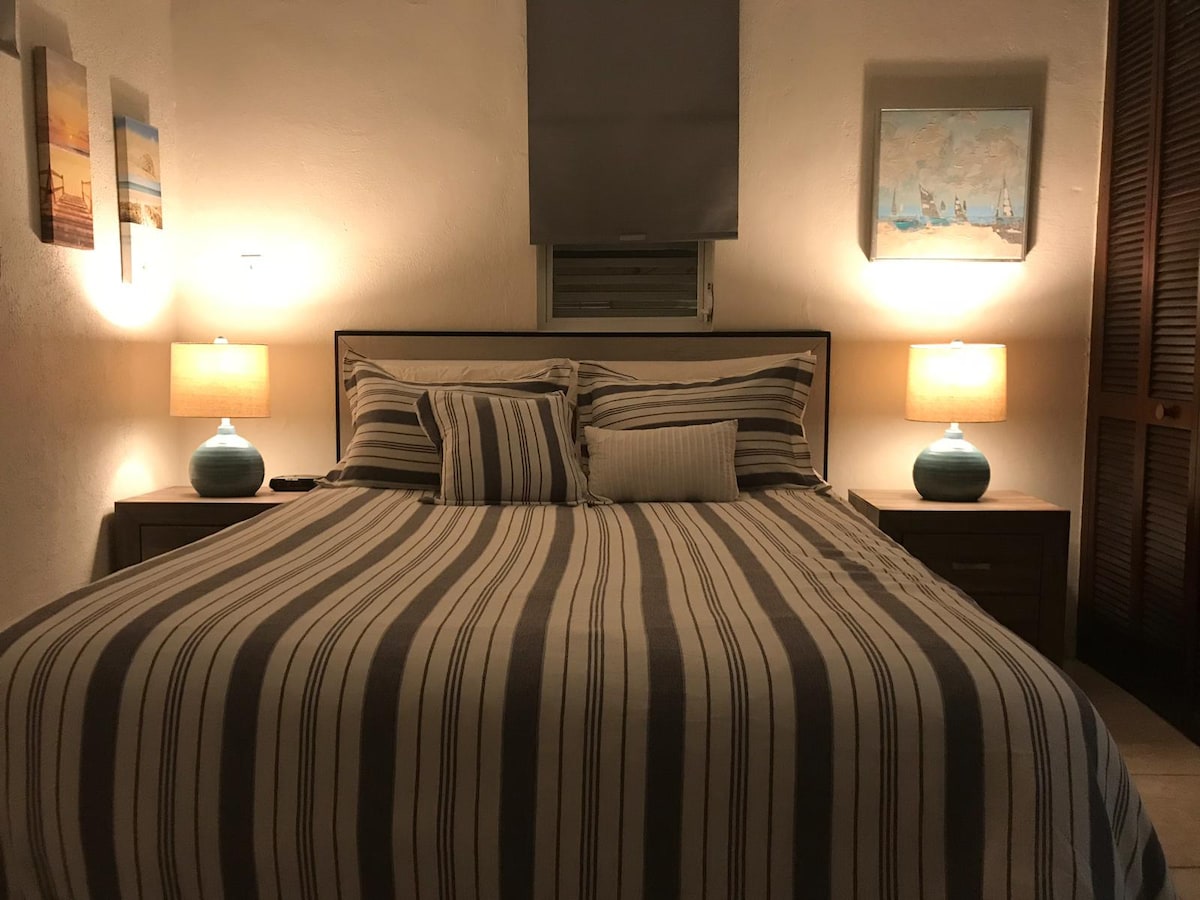 A spacious bed is centered against a wall, adorned with striped bedding and accent pillows. On either side, wooden nightstands hold table lamps with soft light, creating a cozy ambiance. Art pieces are displayed on the walls, adding splashes of color to the room.