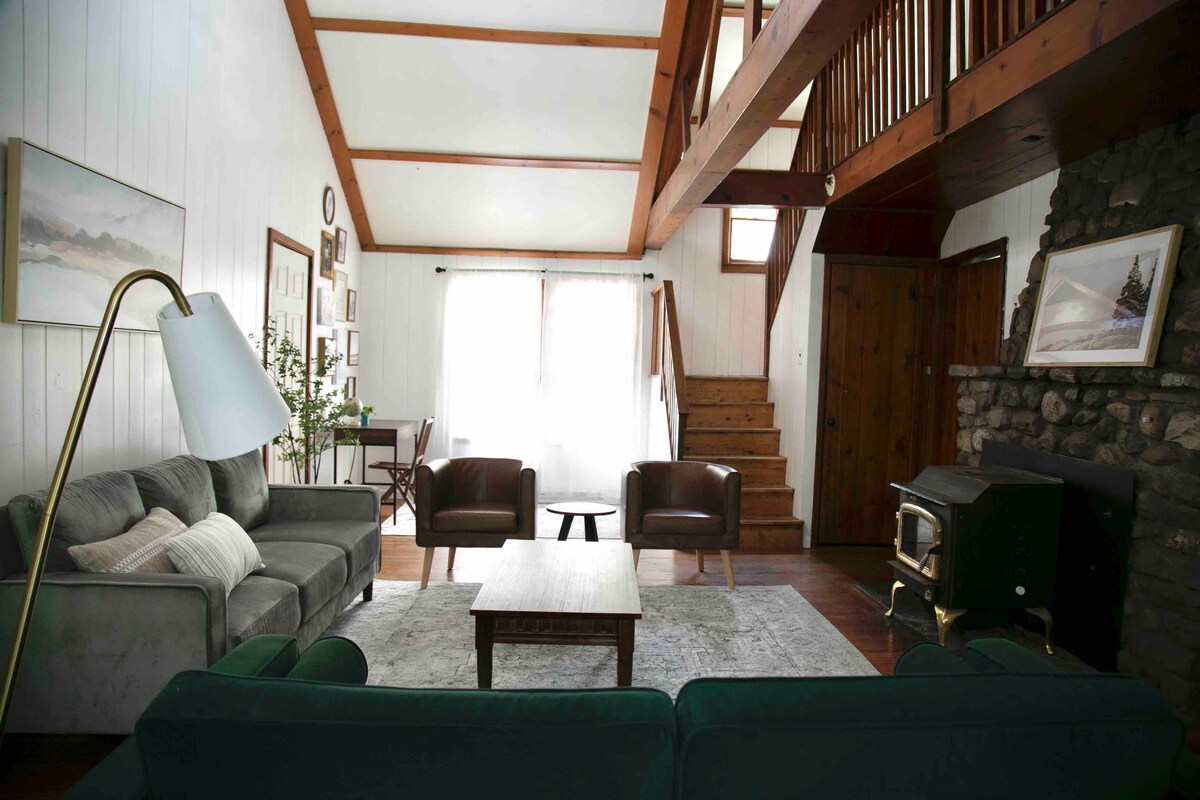 A spacious living area is featured, with two green sofas and two leather armchairs arranged around a wooden coffee table. A stone fireplace stands against one wall, while large windows allow natural light to fill the room. A wooden staircase leads to the upper level.