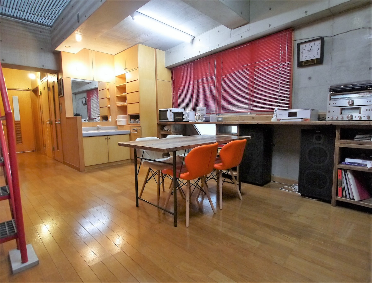 A well-lit shared space is shown with wooden flooring. A wooden table with two orange chairs is located in the foreground. Storage shelves and audio equipment are visible along the walls, alongside a large mirror and sink area. Red curtains provide a touch of warmth.