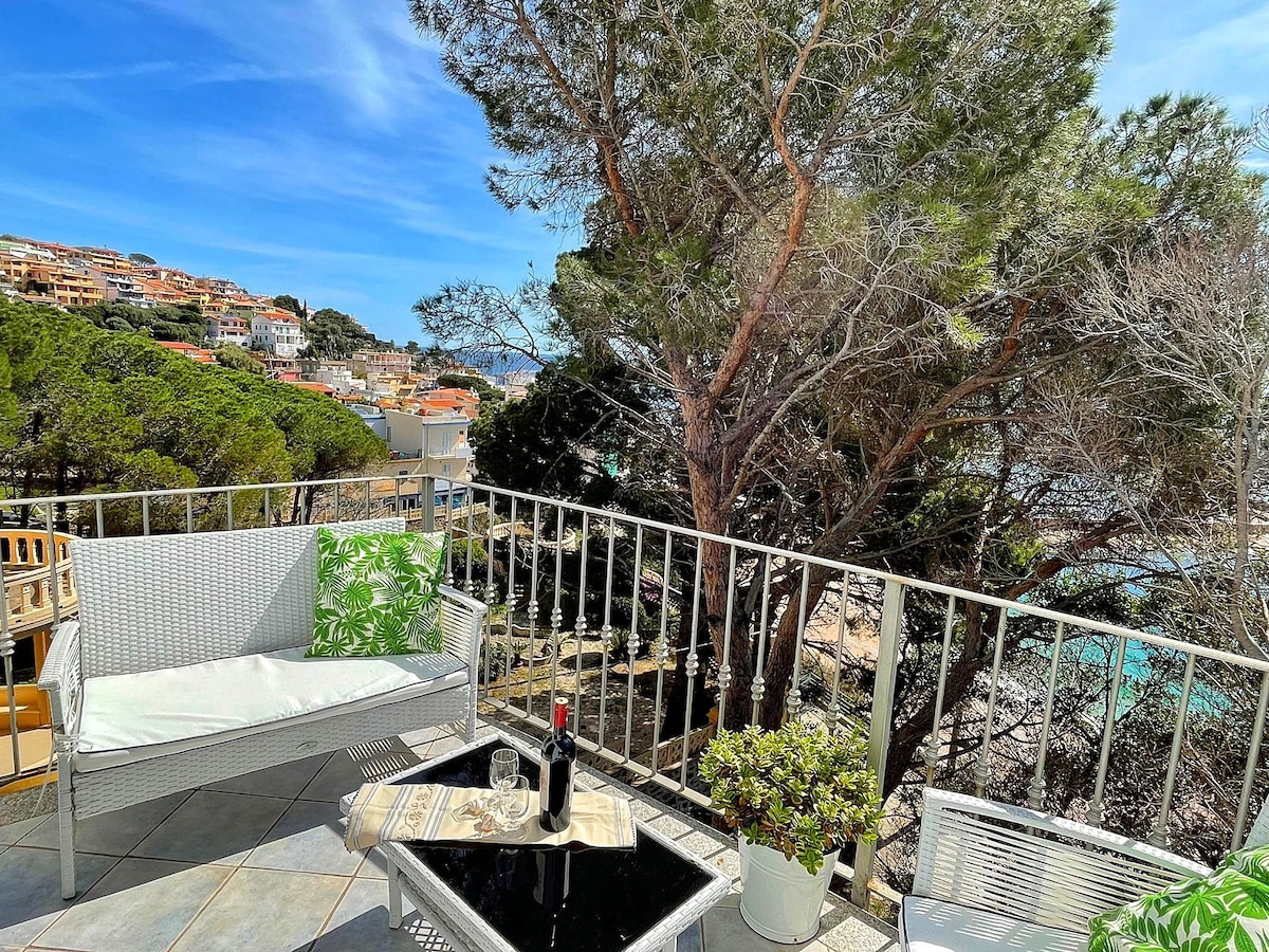 A terrace area is presented with modern outdoor seating, featuring a low black table. Surrounding greenery creates a natural barrier, while views of the coastline and nearby buildings enhance the outdoor setting. A bottle and glass are placed on the table, inviting relaxation.