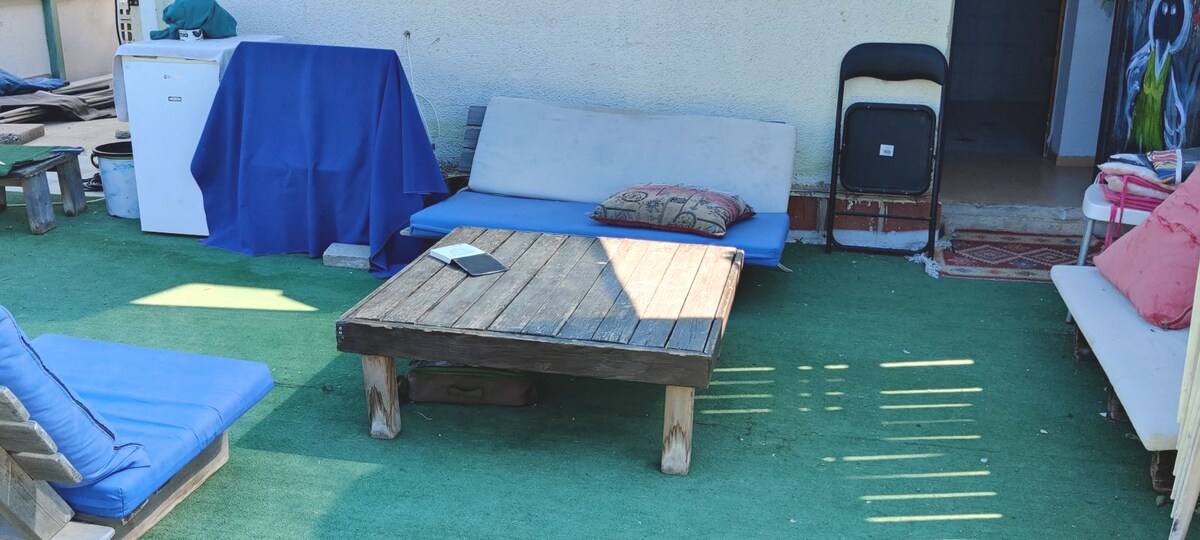 A spacious outdoor area features a wooden coffee table surrounded by lounge seating on a green turf surface. A blue draped item and a small side table are also present, contributing to the relaxed environment. The entrance to the indoor space is visible at the back.