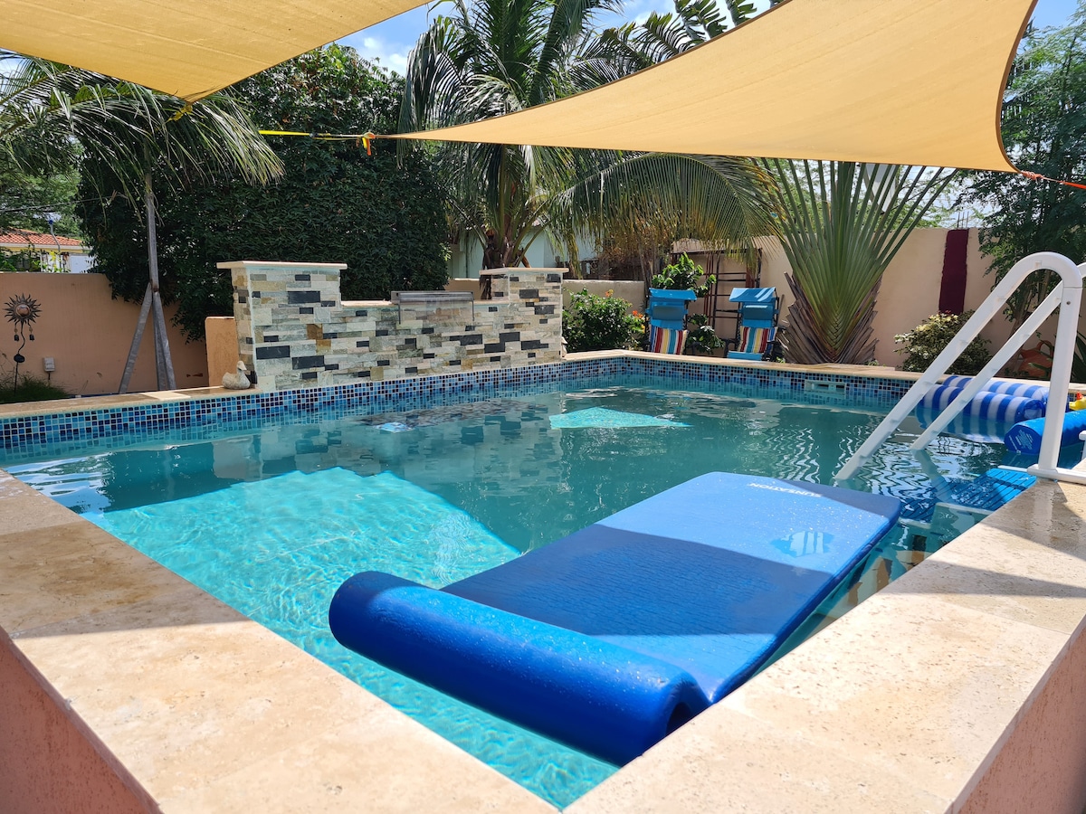 A refreshing pool area is surrounded by sun protection shading and lush greenery. The pool features a smooth entrance with steps, inviting relaxation. A colorful floating lounge is visible in the water, adding to the serene atmosphere of the outdoor space.