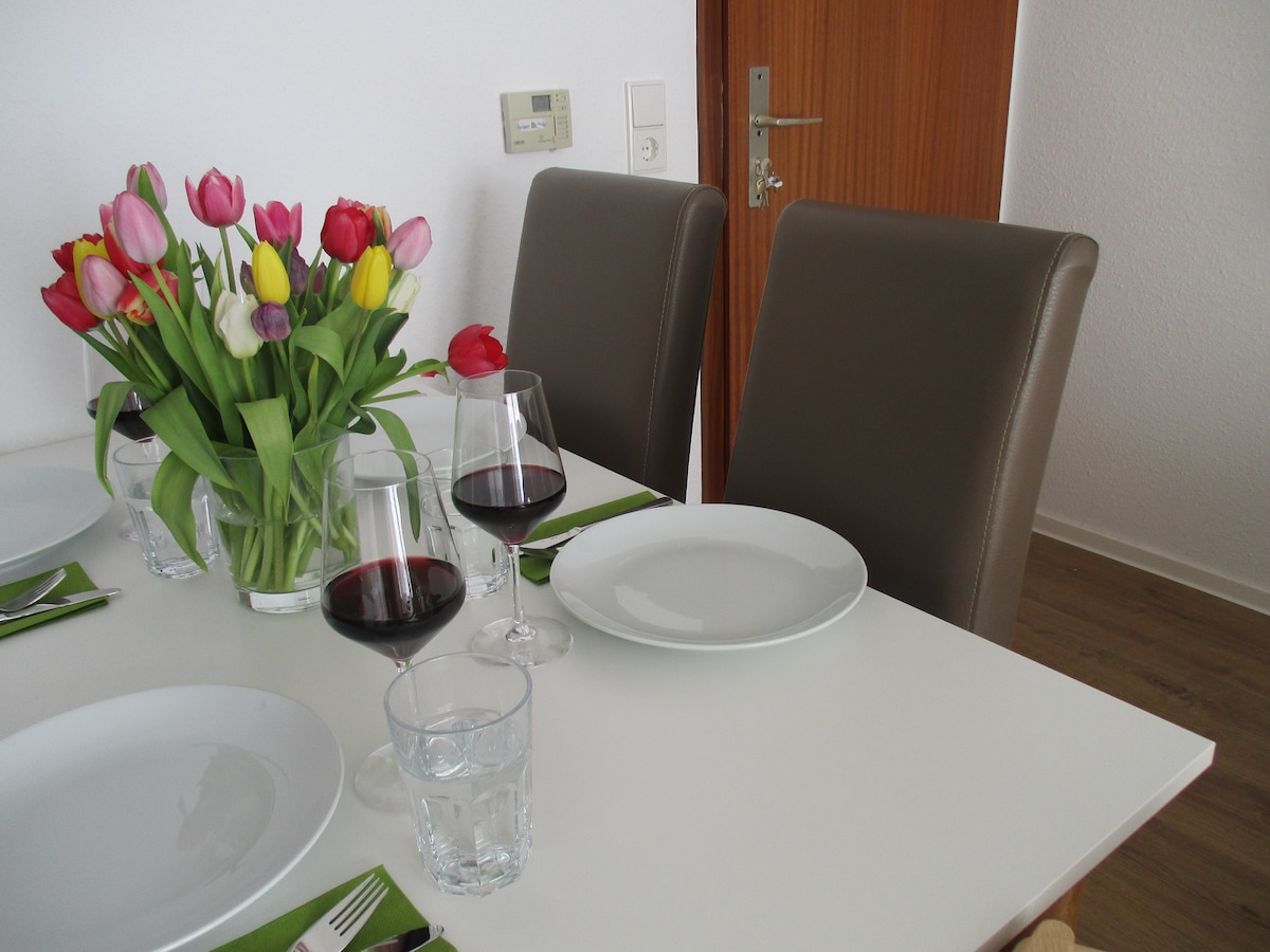 A dining table is set for a meal, featuring round white plates, glasses for water, and wine glasses filled with red wine. A vase of colorful tulips adds a vibrant touch to the table. Light wood flooring complements the modern, neutral-toned chairs.