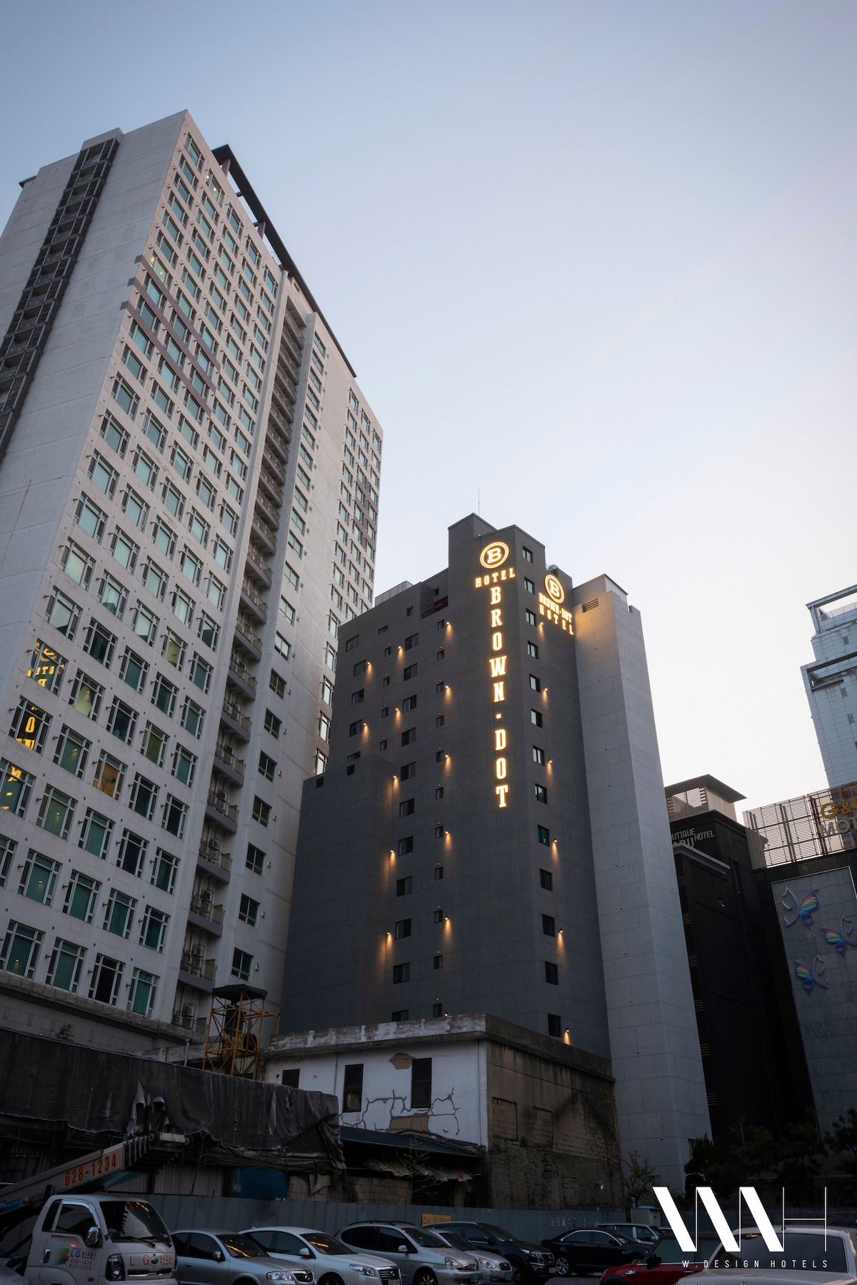 The exterior of Brown Dot Hotel is showcased, emphasizing a tall, modern building with a sleek gray facade. The hotel name is illuminated in bold letters, standing out against the backdrop of the urban skyline during twilight.