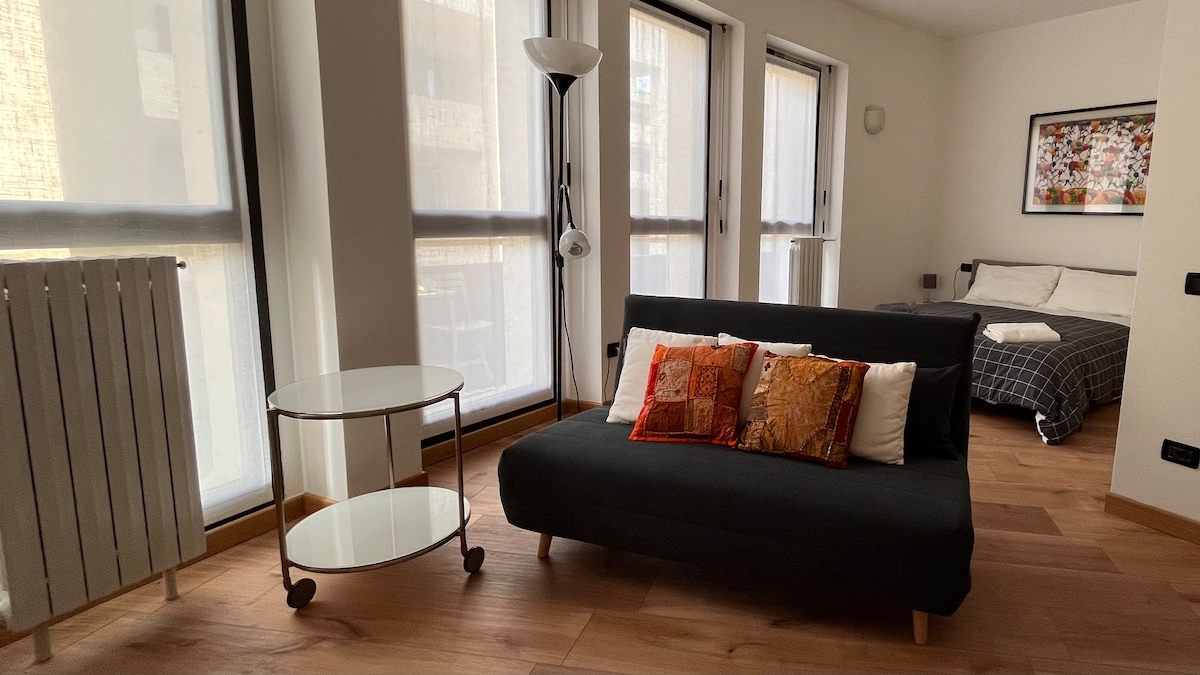 A spacious living area is presented with a dark sofa adorned with colorful cushions. A round glass table complements the seating. Large windows allow natural light to fill the room, with a cozy sleeping area visible in the background featuring a patterned bedspread.