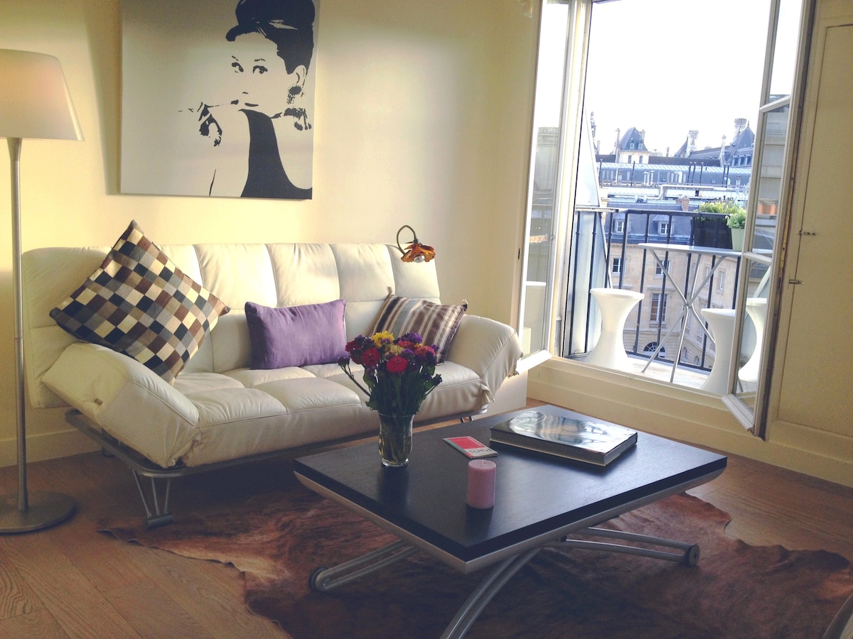 A bright living area features a modern white sofa with decorative cushions and a small coffee table. A bouquet of flowers sits atop the table. Natural light floods through the open balcony doors, providing a view of the Parisian rooftops.