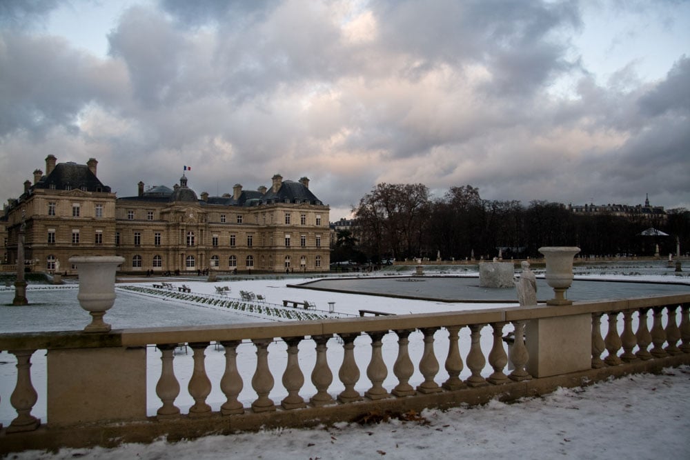 A historic building with classic architectural features is seen behind a snow-covered landscape. The image captures a stone railing in the foreground, leading to a garden area with neatly arranged chairs and symmetrical paths, all set against a cloudy sky.