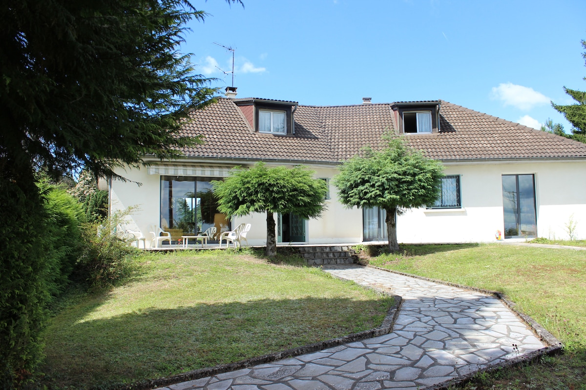 The exterior of a two-story house is showcased, featuring a stone pathway leading to the entrance. The facade is complemented by large glass doors, flanked by greenery. The roof is covered with brown tiles, and windows are partially shaded by small trees.