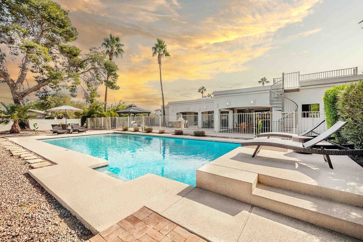 A spacious pool area features a large, inviting pool surrounded by well-manicured landscaping. Comfortable loungers are positioned alongside the water, with a gazebo visible in the background. A sunset sky casts a warm glow over the scene, enhancing the tranquil atmosphere.