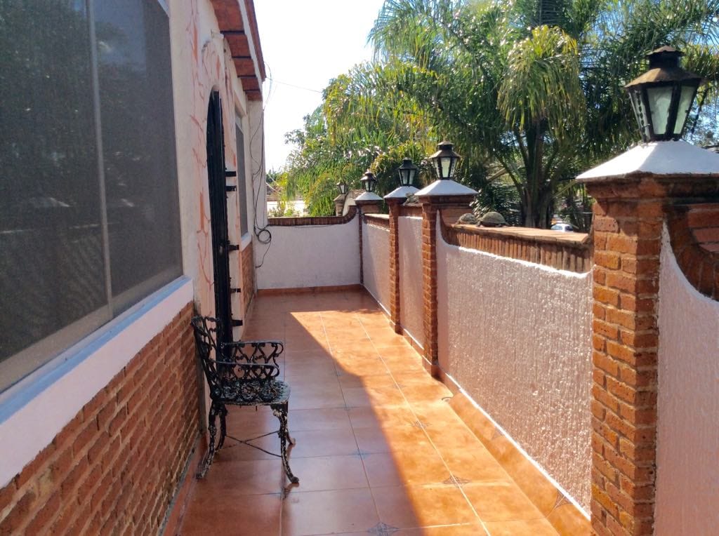 A wide terrace is visible, featuring a terracotta tiled floor and a decorative iron bench positioned against a brick wall. Lined along the space are lantern-style light fixtures, with lush greenery visible in the background, indicating a pleasant outdoor atmosphere.