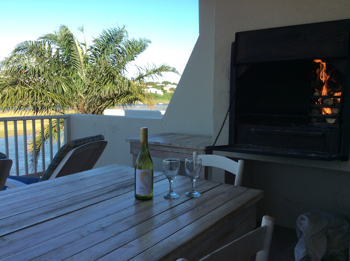 An outdoor dining area is visible, featuring a rustic wooden table accompanied by two glasses and a bottle of wine. A built-in braai is positioned nearby, with flames flickering. Lush palm trees can be seen in the background, framing the serene view of the river.