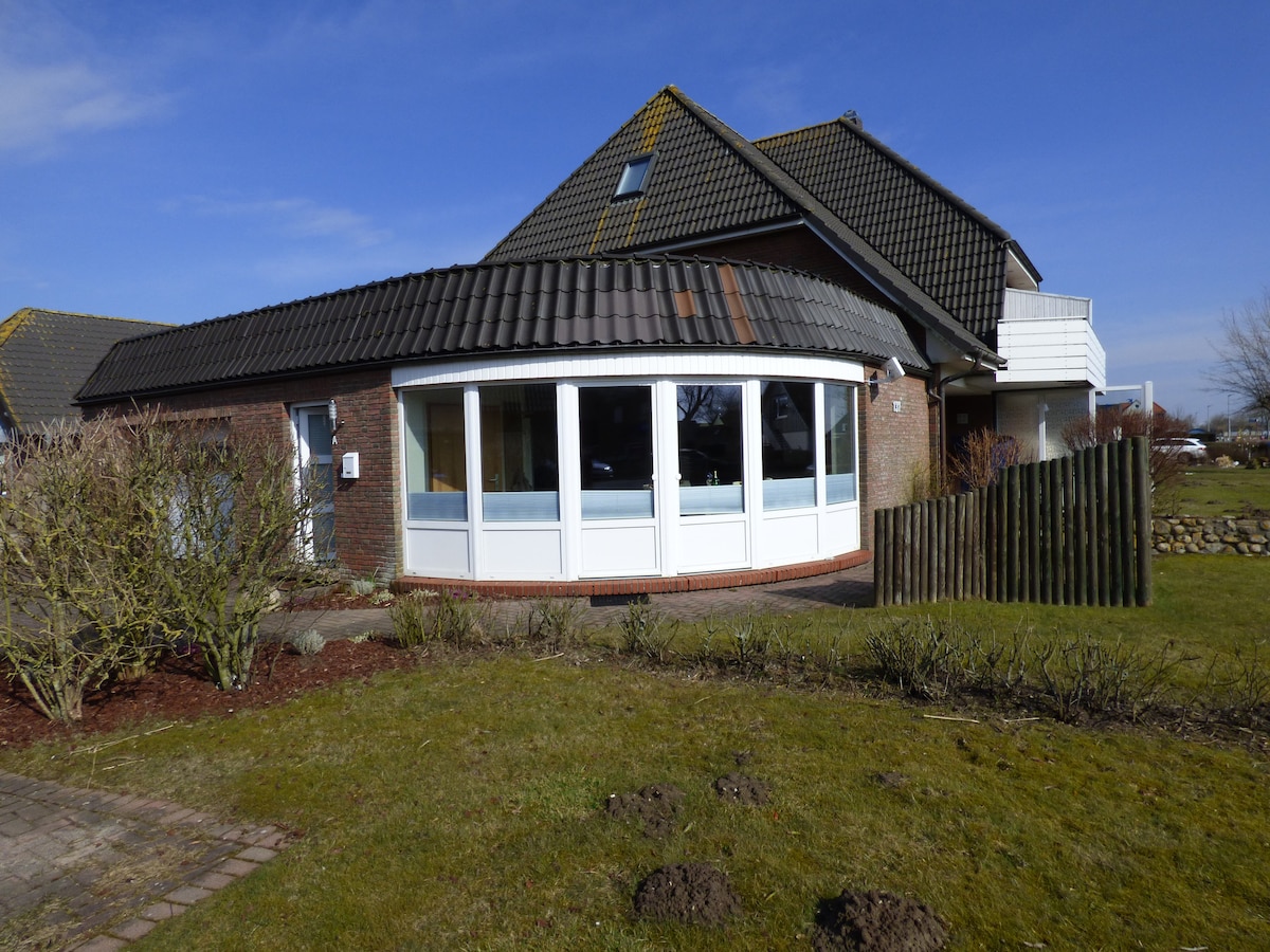 The exterior of the building features a rounded glass section surrounded by a well-maintained garden. A wooden fence and grassy area are visible, along with a mixture of low shrubs. The roof showcases a dark, sloped design against a clear blue sky.