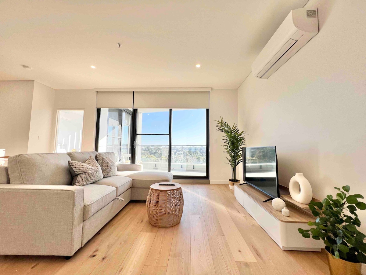 A bright living space features a comfortable light-colored sofa positioned next to a wooden side table. A TV is located on a low white media unit, complemented by a modern decorative piece. Large glass doors offer views of the outdoors and abundant natural light.