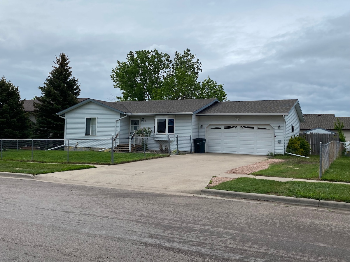 A ranch-style house is shown from the street, featuring a two-car garage and a paved driveway. The front yard has a manicured lawn with a fence surrounding the property. Several trees provide greenery, and clouds hover in the gray sky above.