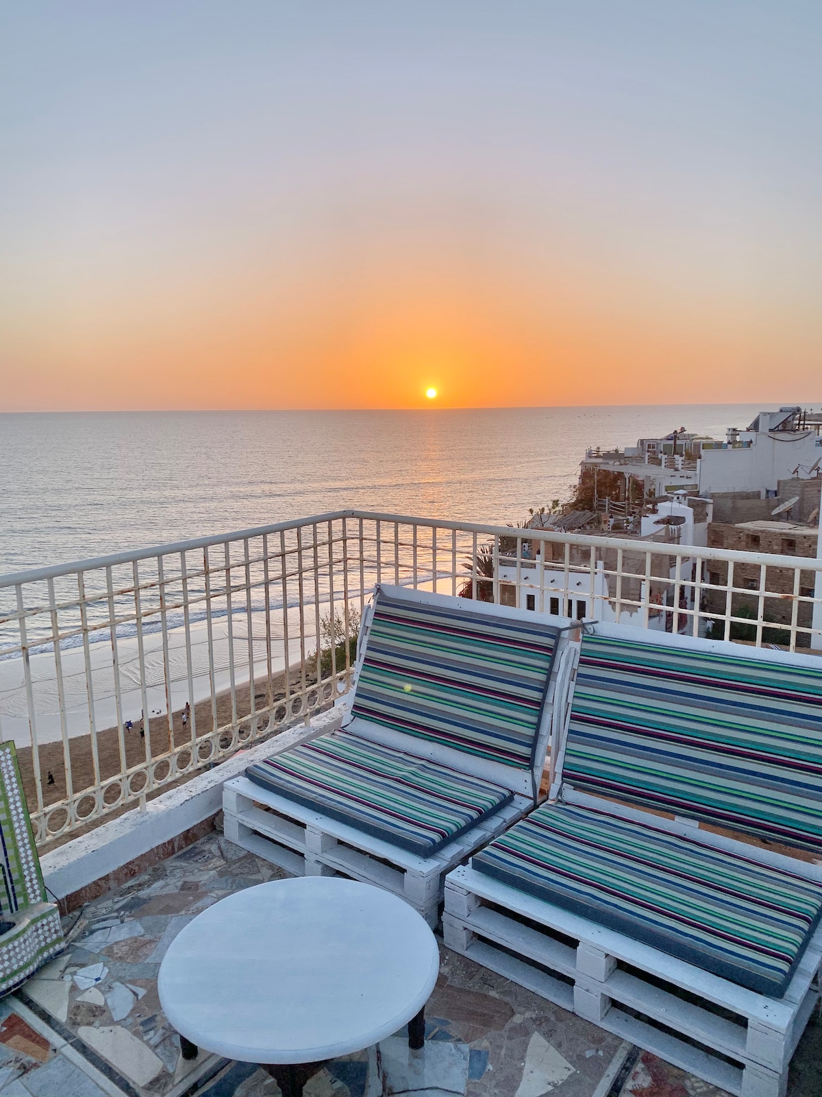 A rooftop terrace offers seating on striped lounge chairs, with a small round table situated between them. A sunset casts warm hues across the sky and reflects on the calm ocean waters, creating a serene coastal atmosphere.