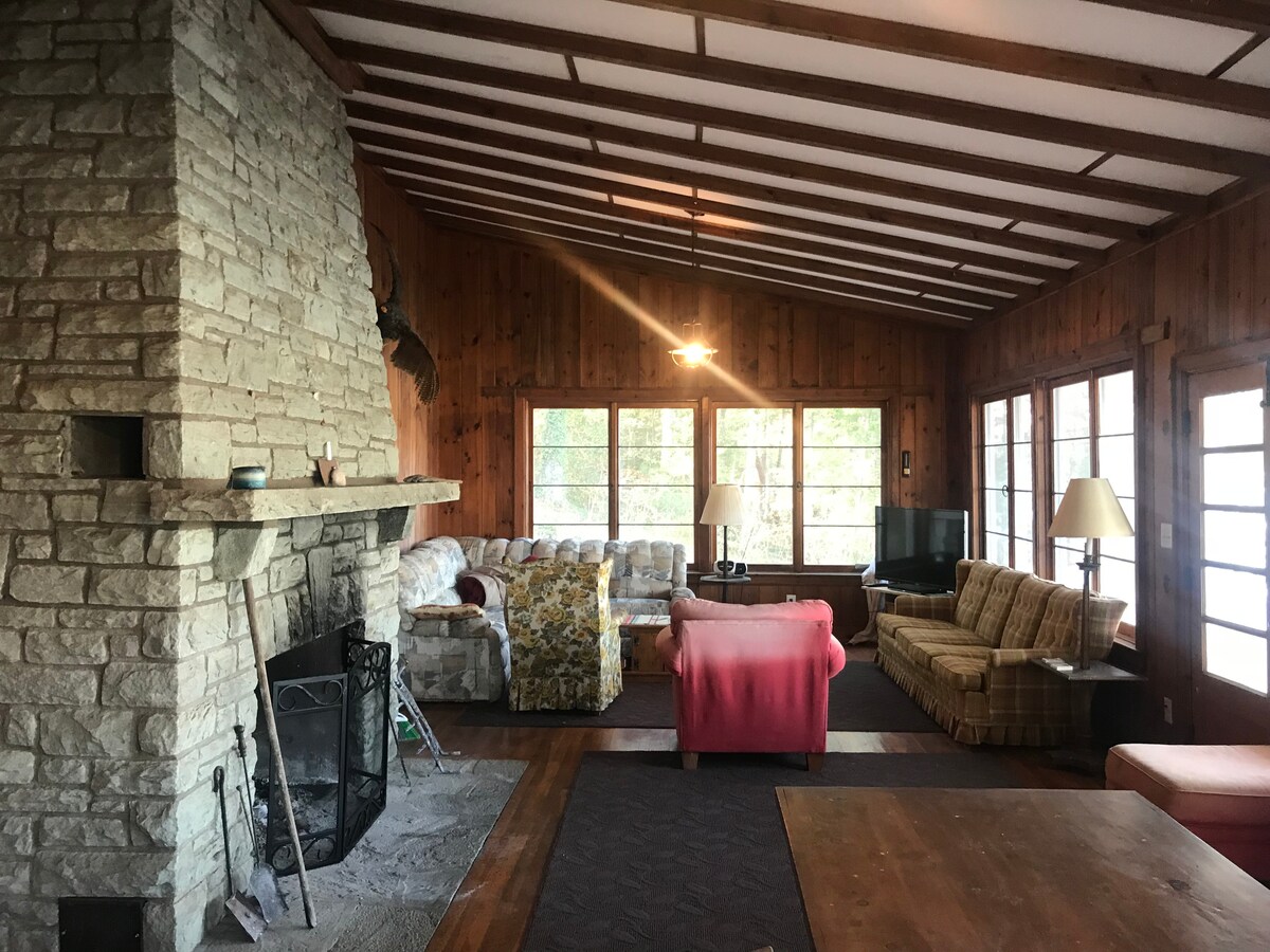 The main living area features a large stone fireplace as a focal point. Comfortable seating options include a floral-patterned sofa and a light-colored sectional. Large windows provide ample natural light and views of the outdoors, while wooden paneling enhances the rustic charm of the space.