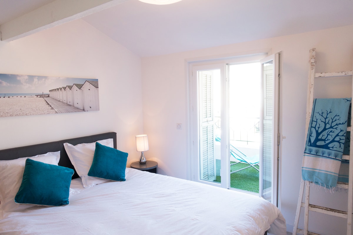 A cozy bedroom features a spacious bed adorned with soft white linens and decorative pillows. Natural light filters in through balcony doors, which open to a private outdoor space. A bedside lamp and a decorative ladder holding a towel enhance the inviting ambiance.