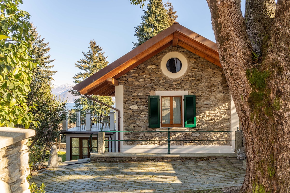 A charming stone cottage is presented with a warm wooden roof, surrounded by greenery. The exterior showcases large windows and traditional green shutters. The patio area is visible, offering a space for relaxation, with glimpses of the natural landscape and distant mountains.