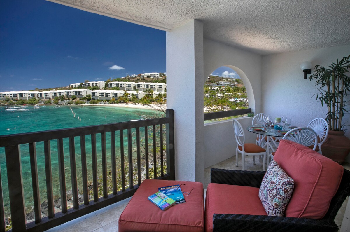 A shaded balcony space features a cozy seating area with orange cushions and a view of the turquoise water. A round dining table is positioned nearby, surrounded by white chairs, with a glimpse of lush landscaping and resort amenities in the background.